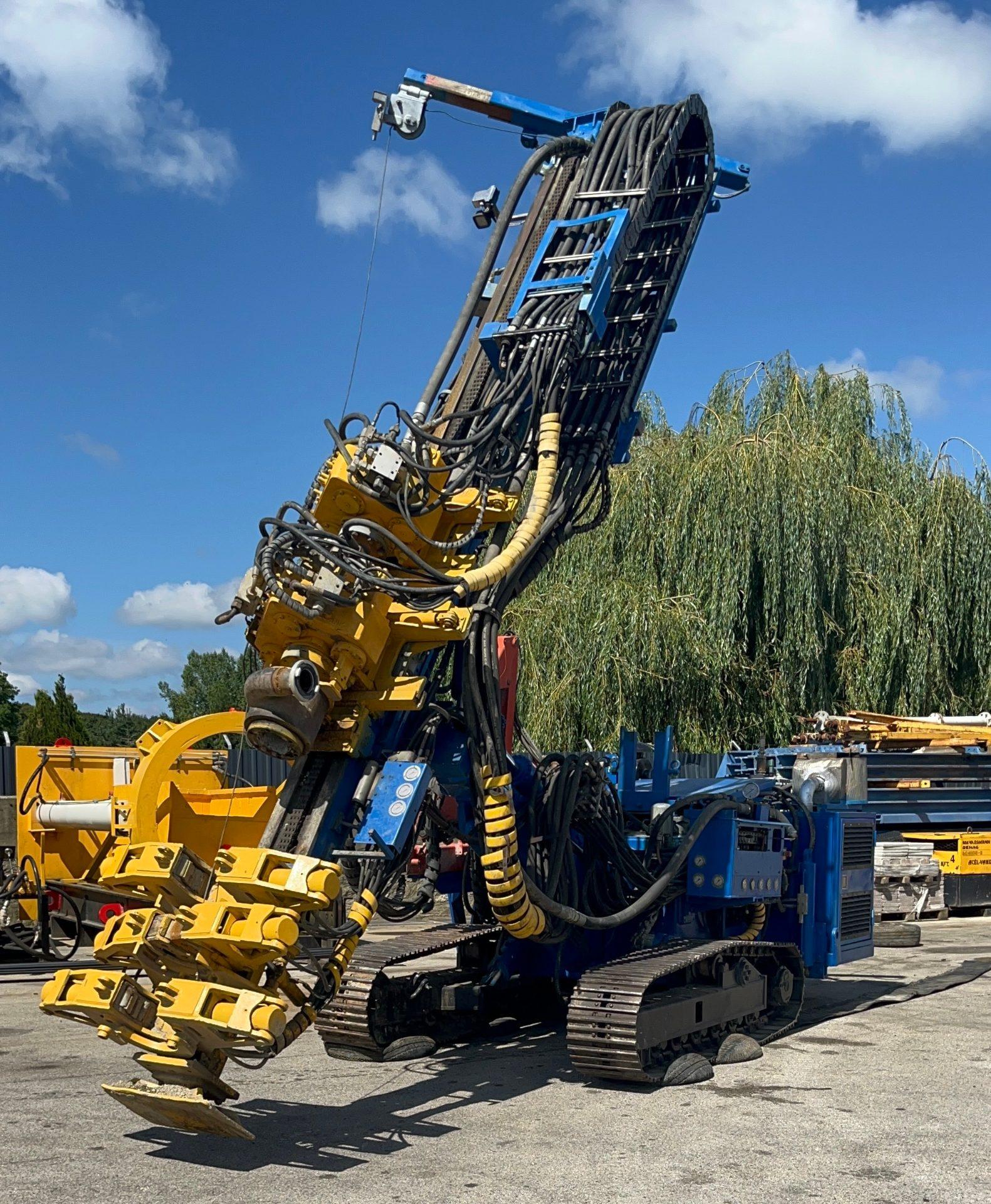 Hütte HBR 504 GT tracked drilling rig, blue and yellow, with a prominent mast, hydraulic hoses, and foundation tools.
